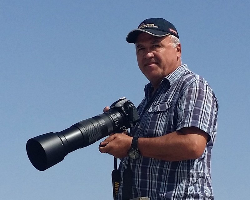 George Yiallouris holding a telephoto camera outdoors against a clear blue sky.